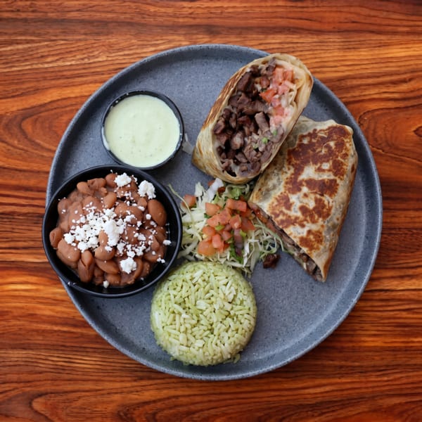 Two burritos, one sliced open, with a side of beans, rice, salad, and green sauce on a plate.